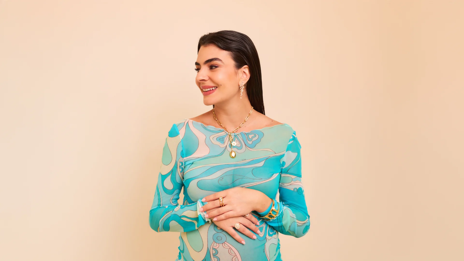 Woman wearing a teal patterned long-sleeve dress, smiling and looking to the side against a neutral beige background, with gold jewelry accents.